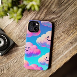 Puede incluir: Una funda de teléfono inteligente con un diseño de nubes alegres. La funda tiene un fondo azul con nubes rosas y blancas sonrientes. La funda del teléfono está sobre una superficie de madera.