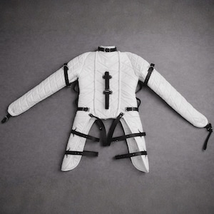 Puede incluir: Chaqueta de sujeción acolchada blanca con correas y hebillas de cuero negro. La chaqueta tiene mangas largas y un cuello. La prenda está diseñada para restringir el movimiento. El fondo es gris liso.