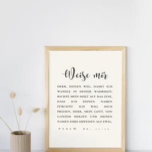 Könnte beinhalten: Ein gerahmter Druck mit dem deutschen Text "Weise mir" in eleganter Schrift. Der Druck zeigt eine religiöse Passage aus Psalm 86:11-12 in schwarzem Text auf cremefarbenem Hintergrund, in einem hellen Holzrahmen. Eine beige Vase mit Trockenblumen befindet sich links.