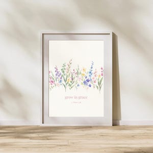 Könnte beinhalten: Ein gerahmter Aquarelldruck mit einem floralen Rand in Lila, Rosa, Gelb und Blau. Der Text "grow in grace" wird in einer zarten rosa Schrift angezeigt. Das Kunstwerk ist vor einem cremefarbenen Hintergrund.