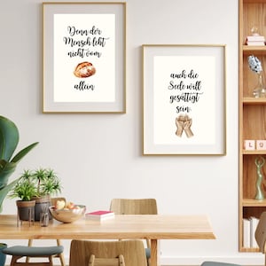 Op de afbeelding: Twee ingelijste kunstprints met gouden frames hangen aan een witte muur. De linkerprint heeft tekst en een aquarel van brood. De rechterprint heeft tekst en een aquarel van handen. Een houten tafel en stoelen staan op de voorgrond.