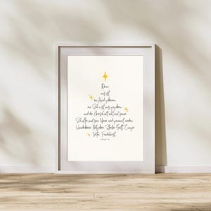 May include: A framed print with a white border, featuring elegant calligraphy in black and gold. The text is arranged in the shape of a Christmas tree, with a star at the top. The print is displayed on a wooden floor.