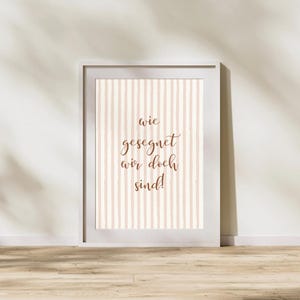 May include: A framed print with a light gray border. The print features a vertical striped pattern in beige and white. The text "wie gesegnet wir doch sind!" is written in brown script.