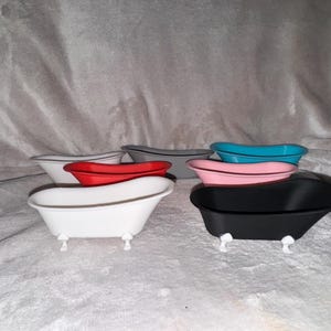 Puede incluir: Una colección de mini bañeras en varios colores: blanco, rojo, gris, rosa, turquesa y negro. Cada bañera tiene pequeñas patas blancas con forma de garra. Las bañeras están dispuestas sobre una superficie suave y texturizada.