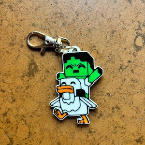 May include: A novelty keychain featuring a green character with black hair riding a white duck with orange accents. The keychain has a silver-colored clasp for easy attachment to bags or keys.
