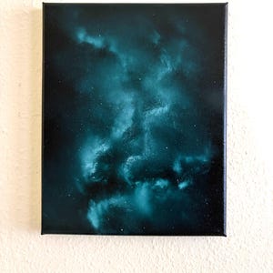 May include: A rectangular canvas painting featuring a deep space nebula theme. The artwork displays swirling clouds of teal and dark blue pigment against a black background, accented with tiny white specks representing distant stars in the galaxy.
