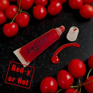May include: A tube of red lip gloss with the text "Lip Lab" and a cherry-shaped applicator. A clear cherry-topped cap and a cherry-shaped design are also present. The phrase "Red-y or Not" is stamped in a red box, surrounded by fresh red cherries.