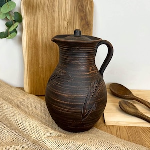 May include: A dark brown ceramic pitcher with a lid and handle. The pitcher features an embossed wheat design and is displayed with wooden spoons and a cutting board. The pitcher is approximately 10 inches tall.