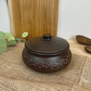 May include: A dark brown ceramic bowl with a lid. The bowl has a decorative floral pattern around the base. The lid has a small knob on top. The bowl is sitting on a burlap surface.