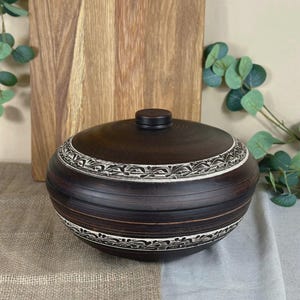 May include: A dark brown, lidded ceramic bowl with a decorative white and black floral pattern around the rim. The bowl sits on a neutral-toned surface, with a wooden cutting board in the background. The lid has a small, round handle.