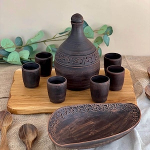 May include: A rustic, dark brown ceramic serving set. The set includes a decorative bottle with a stopper, six small cups, and an oval serving dish. The bottle and dish feature ornate, carved floral patterns. The items are arranged on a wooden board.