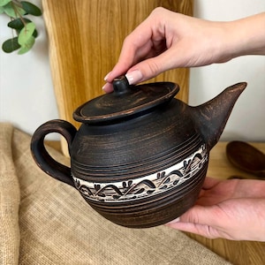 Peut inclure: Une théière en céramique marron foncé avec un couvercle, une anse et un bec verseur. La théière a une bande décorative blanche et marron au milieu. La théière est tenue par une personne.