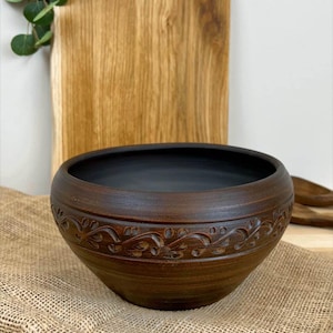 May include: A dark brown ceramic bowl with a black interior. The bowl features a decorative floral pattern around the exterior. A wooden cutting board is in the background, and the bowl sits on a burlap surface.