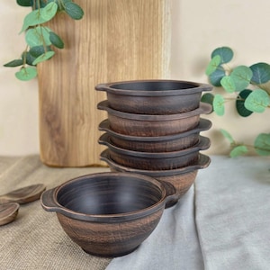 May include: A stack of six rustic brown ceramic bowls with handles. The bowls have a textured surface and a dark brown interior. A wooden cutting board and decorative greenery are in the background.