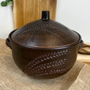 May include: A brown ceramic casserole dish with a lid. The dish and lid feature an embossed wheat stalk design. The dish has two handles and sits on a burlap surface. The background includes a wooden chopping board.