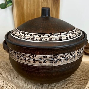 May include: A dark brown ceramic serving bowl with a matching lid. The bowl and lid feature a white band with a repeating leaf design. The bowl has two handles and a small knob on the lid.