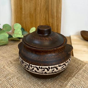 May include: A dark brown ceramic pot with a lid. The pot features a decorative band of white and black floral designs. The pot is on a burlap surface.