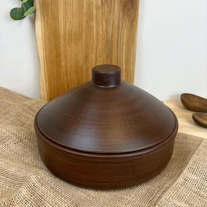 May include: A brown ceramic bowl with a matching lid. The bowl has a rounded shape and a small, cylindrical handle on top of the lid. The bowl is sitting on a burlap cloth.