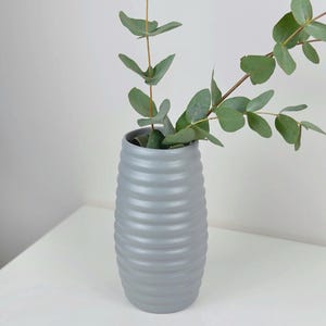 Könnte beinhalten: Eine graue Keramikvase mit geripptem Design hält grüne Eukalyptuszweige. Die Vase ist zylindrisch und steht auf einer weißen Oberfläche. Der Eukalyptus hat mehrere grüne Blätter.