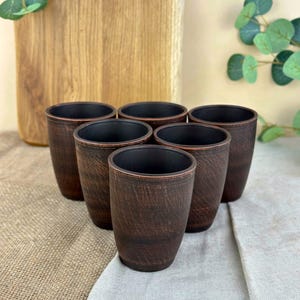 May include: Six brown ceramic cups with a textured finish are arranged in a semi-circle. The cups have a dark interior and are set on a neutral-toned fabric surface. A wooden cutting board is in the background.