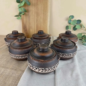 Peut inclure: Six bols en céramique faits à la main, brun foncé, avec couvercles. Chaque bol présente un motif floral blanc décoratif autour de la base. Les bols sont disposés sur une surface claire, avec une planche à découper en bois et de la verdure en arrière-plan.