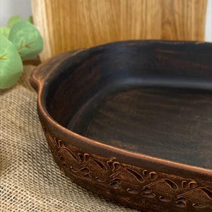 May include: A dark brown, oval-shaped ceramic baking dish with a decorative floral pattern around the rim. The dish has a rustic, handmade appearance and is suitable for baking and serving. The interior is a deep, rich brown color.