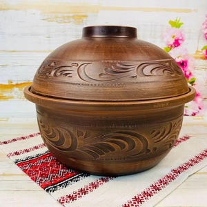 Könnte beinhalten: Ein brauner Keramikkochtopf mit Deckel, mit geschnitzten floralen Designs. Der Topf steht auf einem weiß-rot gemusterten Tuch. Der Hintergrund umfasst eine Holzoberfläche und rosa Blumen.