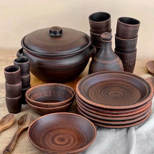 Może przedstawiać: Zestaw rustykalnych brązowych naczyń ceramicznych, w tym duży garnek z pokrywką, talerze, miski, kubki i butelka. Widoczne są również drewniane łyżki. Ceramika ma teksturowane wykończenie i ciepły, ziemisty odcień.