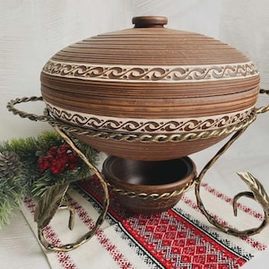 Può includere: Una zuppiera in ceramica marrone con coperchio, su un supporto decorativo in metallo. Il piatto presenta una fascia a motivi bianchi e una ciotola più piccola. Il supporto ha un design a torsione e accenti di foglie.