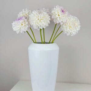 Puede incluir: Un jarrón blanco brillante contiene un ramo de dalias blancas con puntas de color púrpura pálido. Las flores tienen tallos verdes y están dispuestas para crear una exhibición completa y texturizada. El jarrón es cilíndrico y está sobre una superficie blanca.