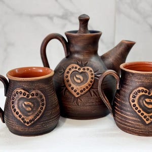May include: A rustic brown ceramic tea set featuring a teapot and two mugs. Each piece has a heart design with a coffee bean in the center, surrounded by small dots. The interior of the mugs is a warm orange color.