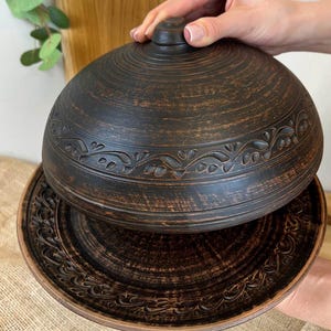 May include: A dark brown ceramic serving dish with a lid. The dish and lid have a textured, rustic appearance with carved floral patterns around the edges. The lid is being lifted, revealing the dish's interior.