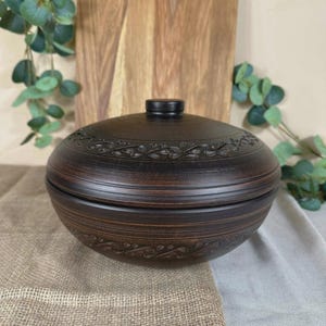 May include: A dark brown ceramic bowl with a lid. The bowl has a rounded shape with decorative etched patterns around the rim and on the lid. The lid has a small, round handle. The bowl is sitting on a burlap and linen surface.