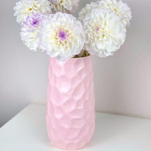 Peut inclure: Un bouquet de dahlias blancs avec des centres violets dans un vase rose clair texturé. Le vase présente un motif géométrique et repose sur une surface blanche. Les fleurs sont en pleine floraison.