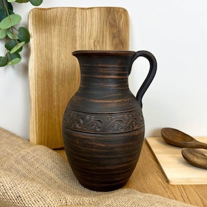 Peut inclure: Une cruche en céramique marron foncé avec une anse et une bande décorative en relief. La cruche est présentée sur une surface en bois avec une planche à découper en bois et du tissu de jute en arrière-plan. Un article de cuisine rustique.