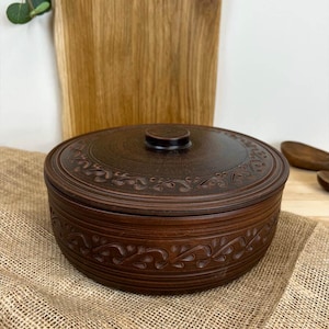 Peut inclure: Un bol en céramique marron avec un couvercle, orné d'un motif floral décoratif sur le bord et le couvercle. Le bol est posé sur une surface en toile de jute, avec une planche à découper en bois en arrière-plan.