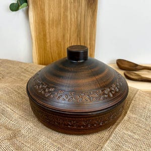 May include: A brown ceramic lidded bowl with a floral pattern around the rim. The bowl has a dark brown top and a lighter brown base. The lid has a small, round handle. The bowl is sitting on a burlap surface.