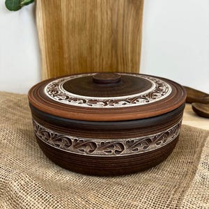 Può includere: Una ciotola in ceramica marrone con coperchio. La ciotola presenta un motivo decorativo bianco e marrone sul bordo e sul coperchio. La ciotola è appoggiata su una superficie di juta.