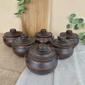 May include: Six small, dark brown ceramic pots with lids are displayed. Each pot has a rounded shape, textured surface, and small handles. The pots are arranged on a neutral-coloured cloth, suitable for a kitchen or dining setting.