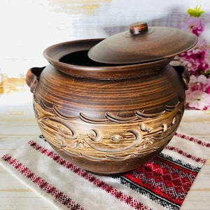 May include: A brown ceramic pot with a lid slightly ajar, featuring carved floral designs around the base. The pot has two handles and sits on a patterned cloth with red and white embroidery. Decorative kitchenware.