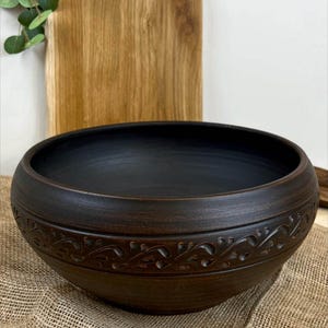 May include: A dark brown ceramic bowl with a wide, round shape. The bowl has a decorative, carved floral pattern around the rim. The interior of the bowl is black. The bowl is sitting on a burlap surface.