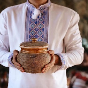 May include: A brown ceramic jar with a lid is held in front of a white shirt with blue and white embroidery. The jar has a woven texture on the bottom and a smooth, light brown top.