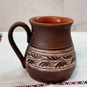 May include: A handmade brown ceramic mug with a curved handle. The mug features a decorative band with a leaf pattern in a lighter shade of brown. The interior of the mug is a reddish-brown color.