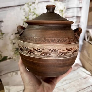 May include: A brown ceramic cooking pot with a lid and two handles. The pot features a decorative band with a leaf design and a lighter-colored band around the middle. The pot is round and appears to be handmade.