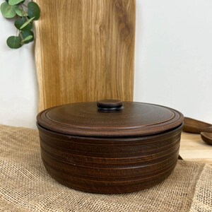 Può includere: Una ciotola in ceramica marrone con coperchio. La ciotola presenta scanalature orizzontali e un colore marrone scuro. Il coperchio ha una piccola maniglia rotonda. Un tagliere in legno è sullo sfondo.