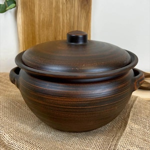 May include: A dark brown ceramic cooking pot with a matching lid. The pot has two small handles and a rounded shape. The lid has a small knob on top. The pot is sitting on a burlap surface.