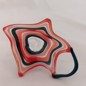 May include: A unique, handmade glass mug with a star-like shape. The mug features a layered design of red, white, and dark teal stripes. The handle is dark teal. The interior of the mug is clear glass.