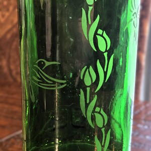May include: A green glass tumbler with etched designs. The tumbler features a hummingbird and tulip pattern in a lighter shade of green. The glass has a slightly textured surface and a dark green hue.