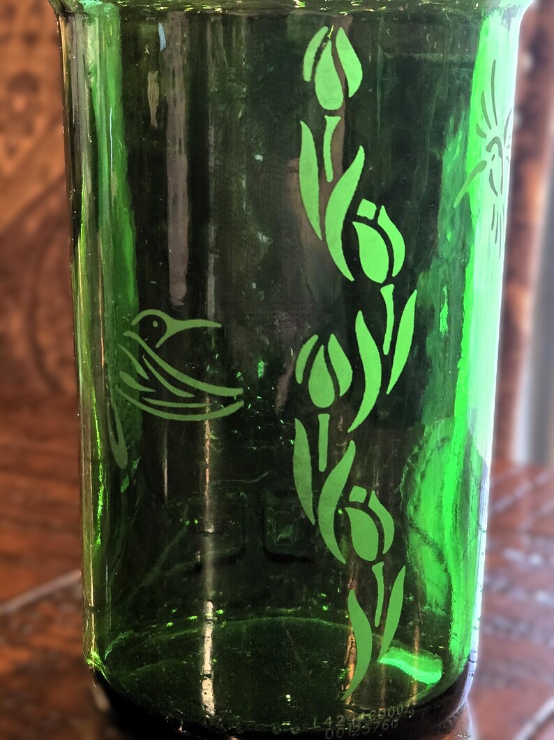 May include: A green glass tumbler with etched designs. The tumbler features a hummingbird and tulip pattern in a lighter shade of green. The glass has a slightly textured surface and a dark green hue.