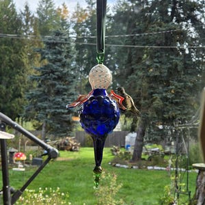 May include: A handcrafted glass hummingbird feeder with a blue, bulbous body and a long, pointed bottom. The feeder has red and orange wings and a white and clear patterned top. A hummingbird is perched on the feeder.